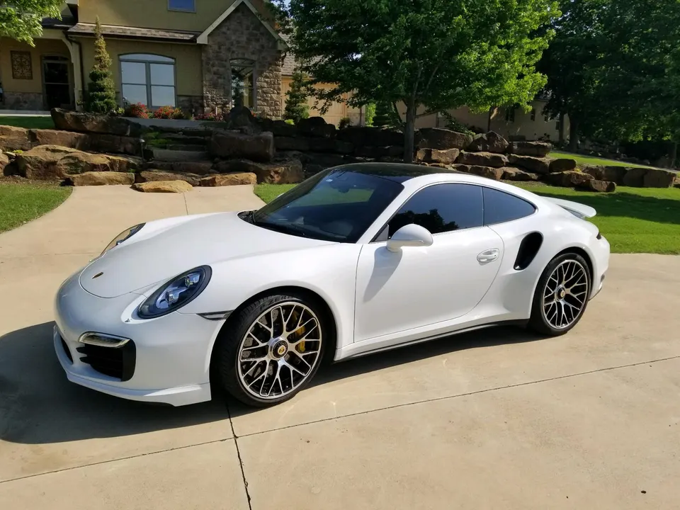 A white Porsche 911 Turbo S parked on a sunny driveway in front of a large house.