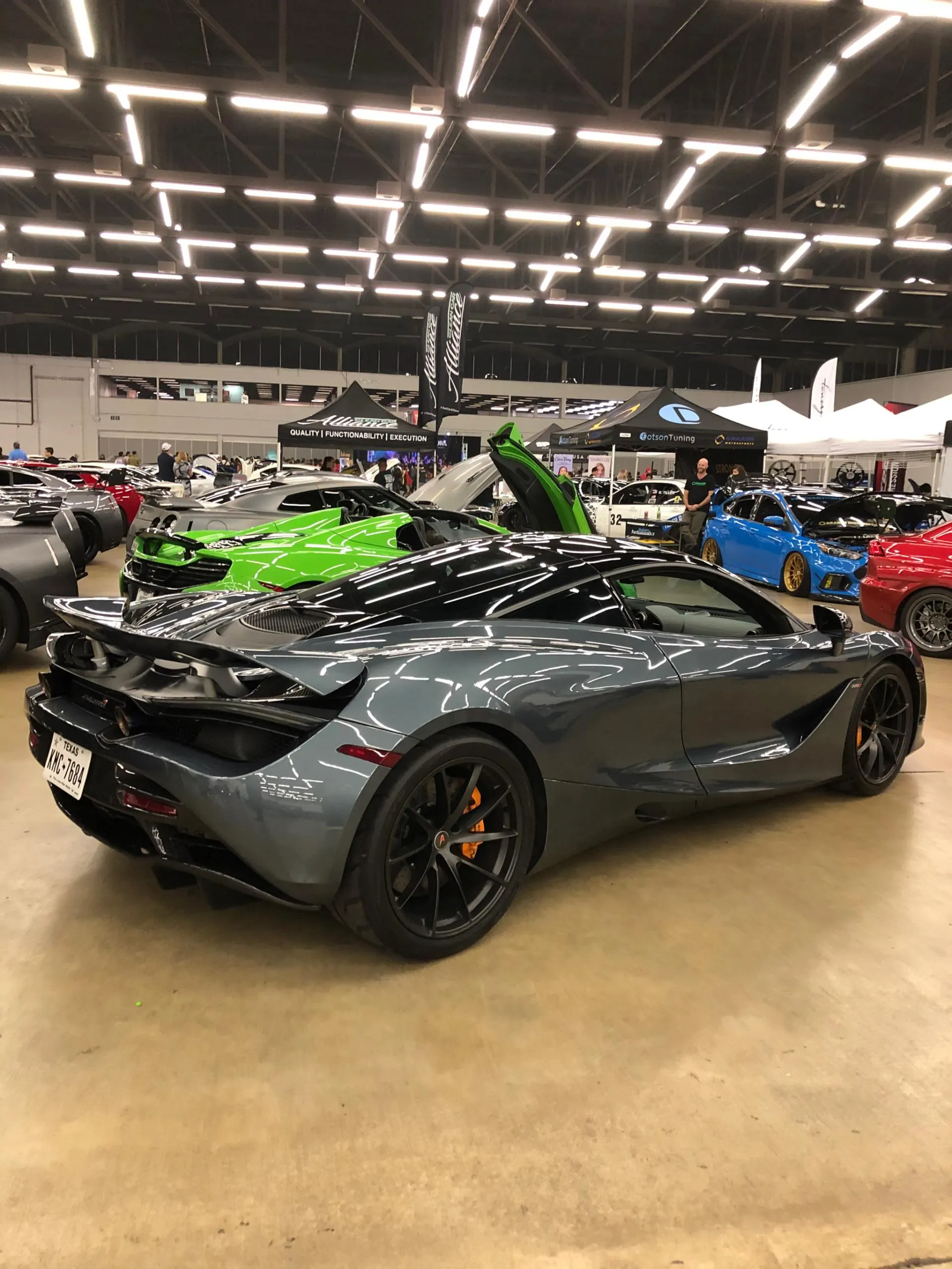 Grey McLaren 720S parked at a car show with other performance vehicles and attendees in the background.