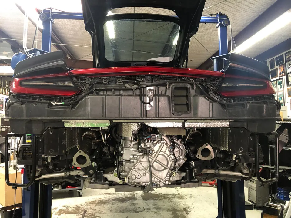 Rear view of a Corvette C8 on a lift in a shop with the bumper removed, exposing the engine and exhaust system.