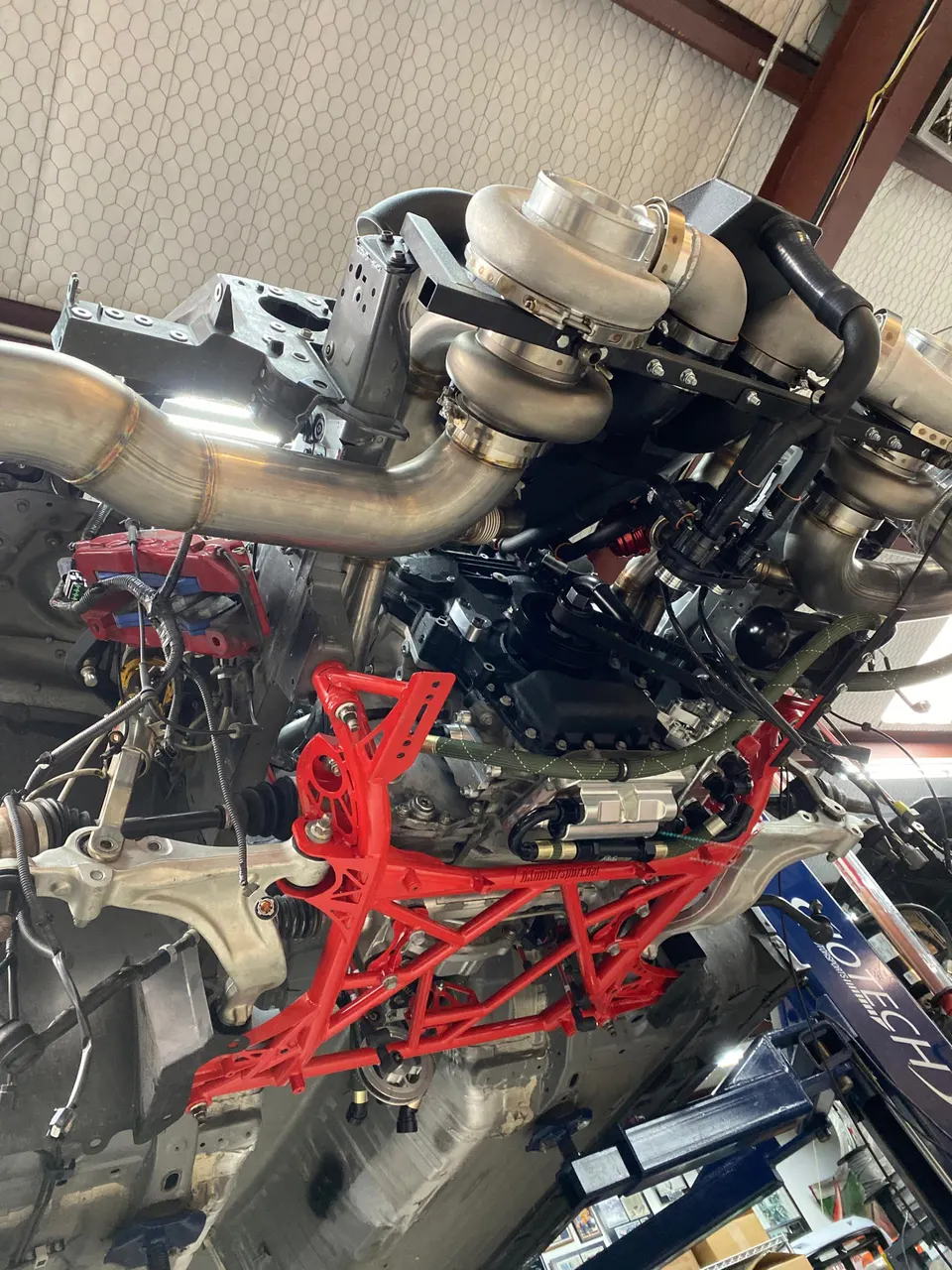 Underside view of a high-performance car on a lift, showcasing twin turbochargers, custom exhaust, and a red tubular subframe.