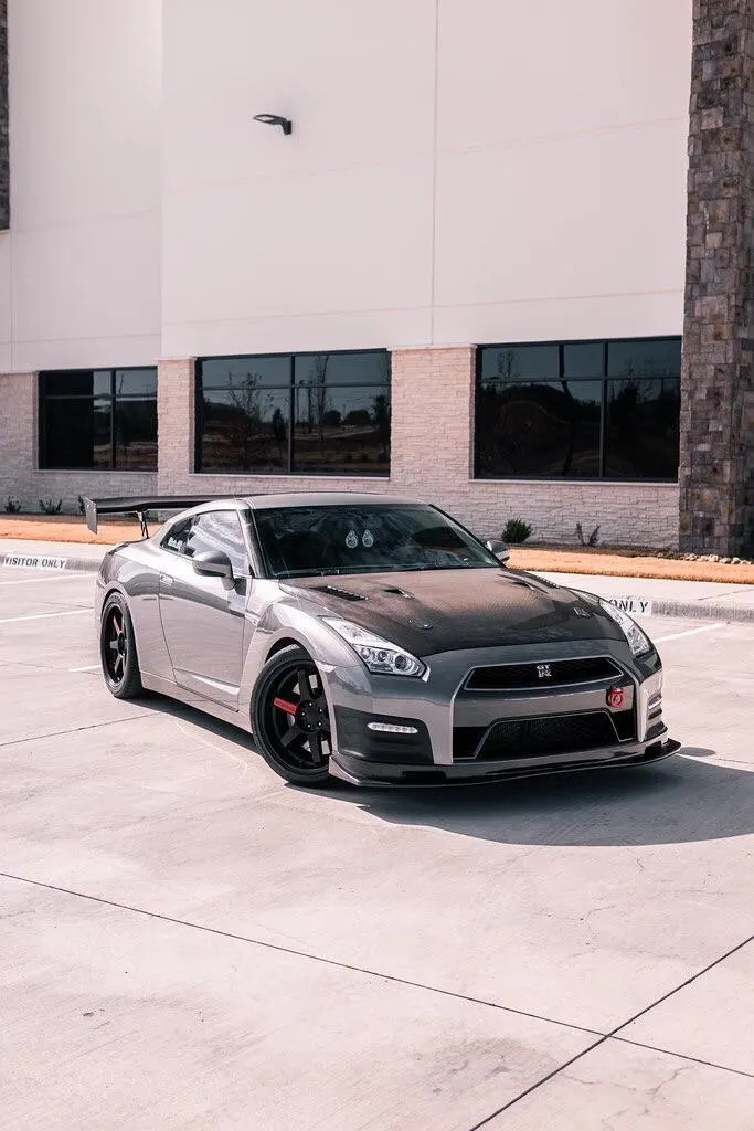 A grey Nissan GT-R with a carbon fiber hood, aftermarket wheels, and a large rear wing parked in front of a modern building.