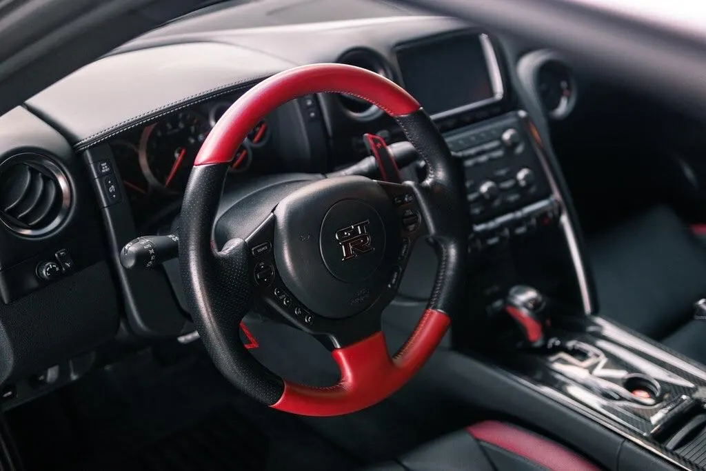 A close-up of a Nissan GT-R interior, featuring a black and red steering wheel with paddle shifters and dashboard.