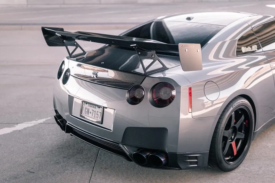 A silver Nissan GT-R with a large carbon fiber wing, carbon fiber trunk, and aftermarket exhaust system parked on a street.