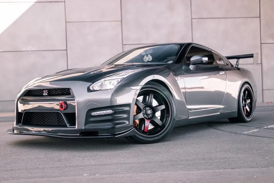 A grey Nissan GT-R R35 with aftermarket wheels, carbon fiber hood, and front splitter parked in front of a modern building.