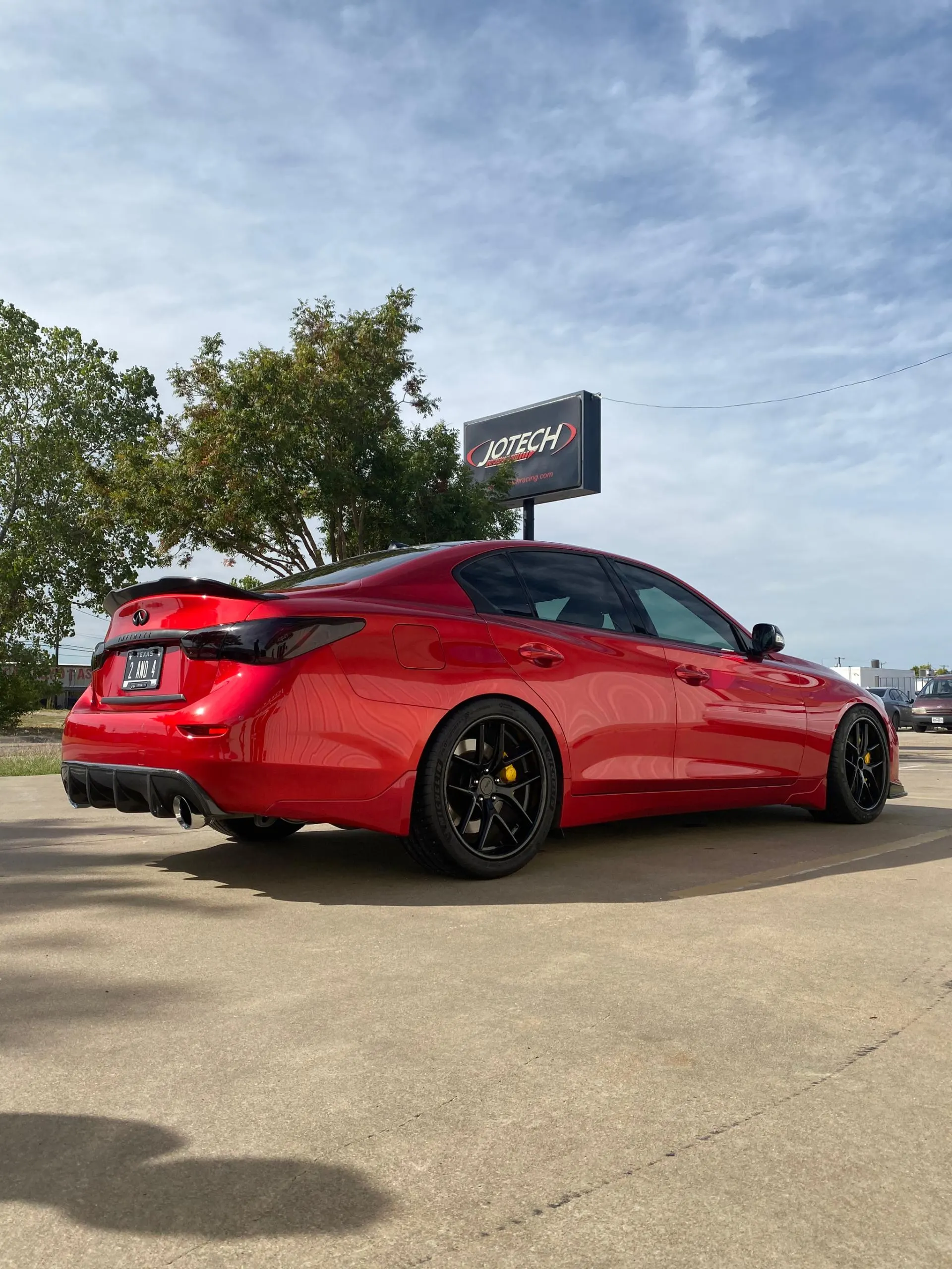 Red Infiniti Q50 with aftermarket black wheels, yellow brake calipers, rear diffuser, and trunk spoiler parked outside Jotech Motorsports.