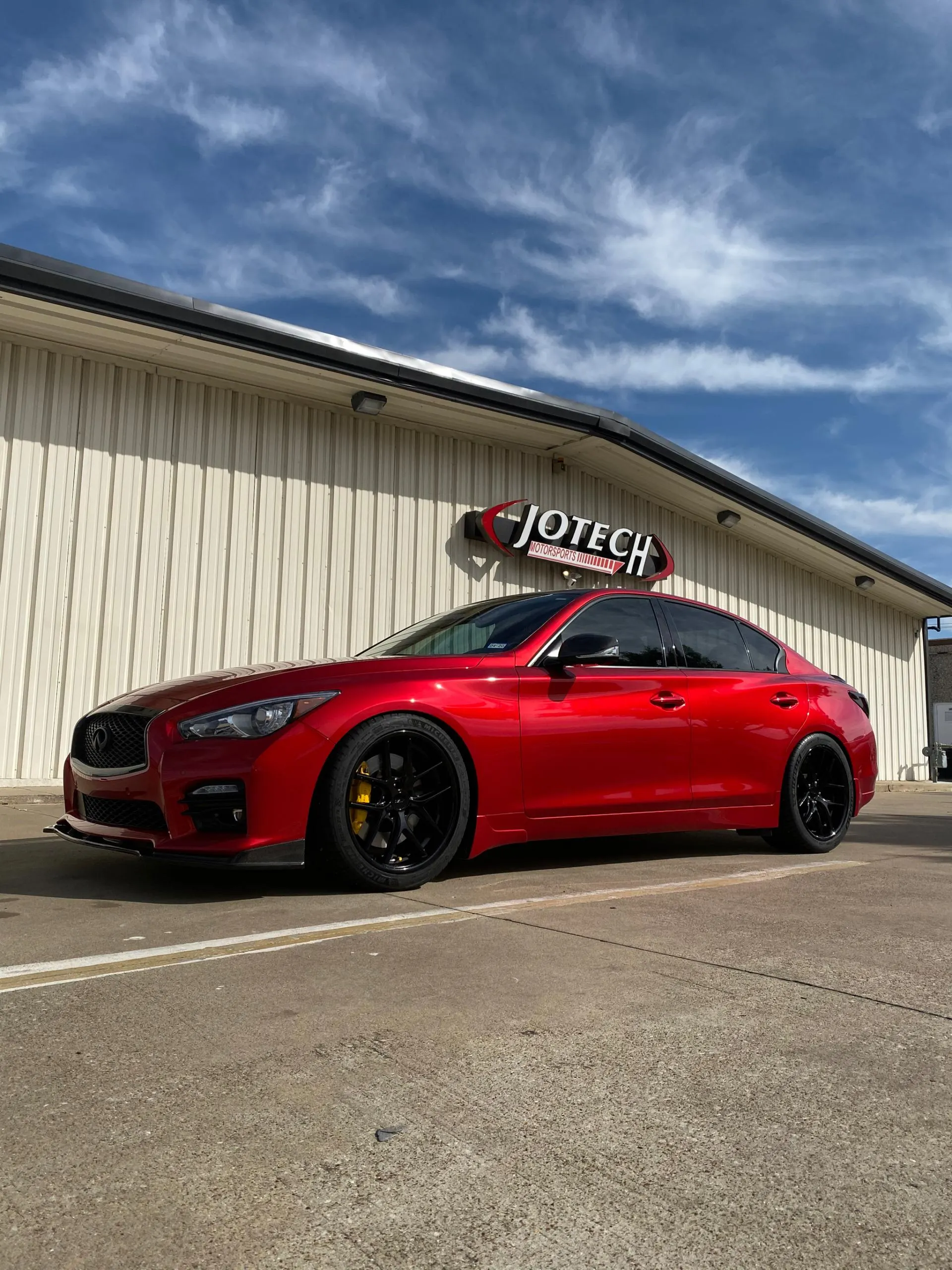A red Infiniti Q50 sedan with aftermarket black wheels and yellow brake calipers parked outside Jotech Motorsports.