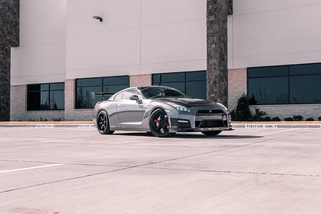 A silver Nissan GT-R with aftermarket wheels, a carbon fiber hood, and a large rear wing parked in front of a modern building.