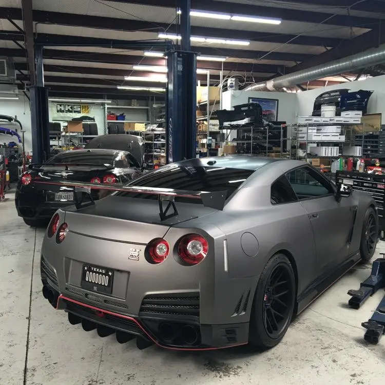 Two modified Nissan GT-Rs, one matte grey and one black, parked inside a performance tuning shop with lifts and parts.