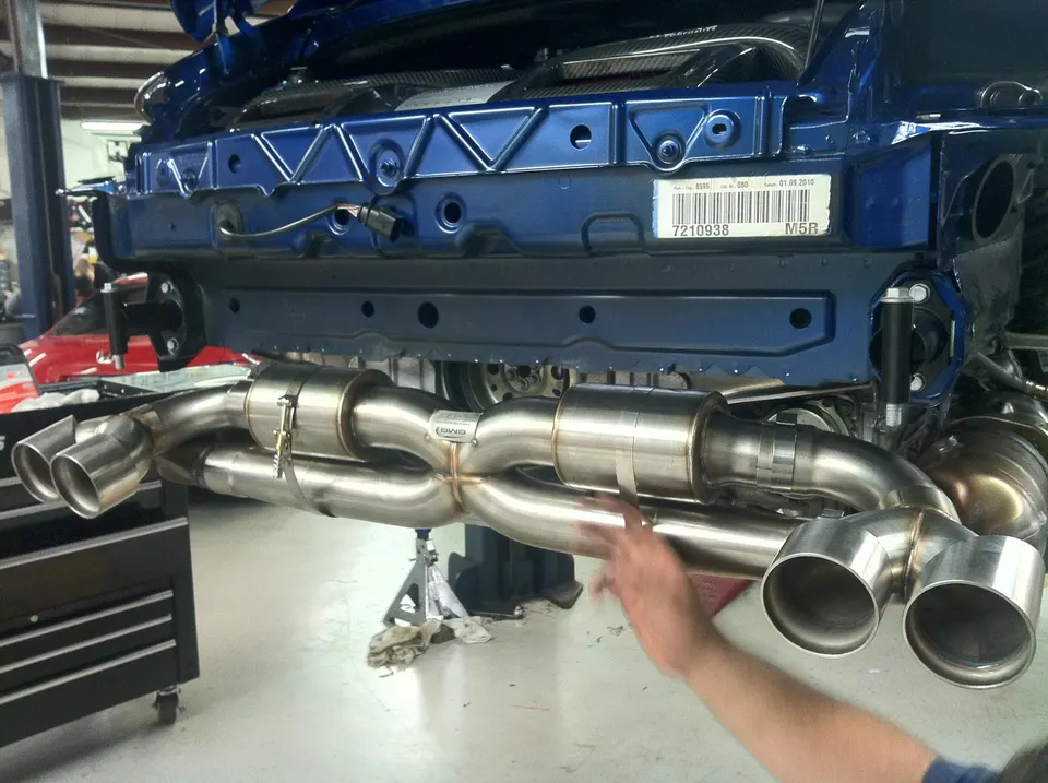 New stainless steel exhaust system being installed on a blue sports car in a performance tuning shop.