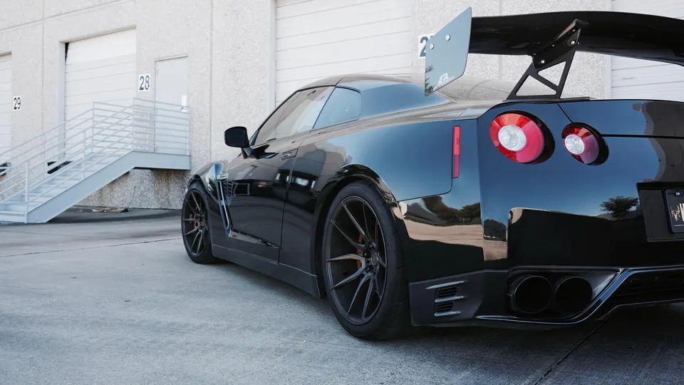 Black Nissan GT-R with a large aftermarket wing, custom wheels, and quad exhaust tips parked outside a building.