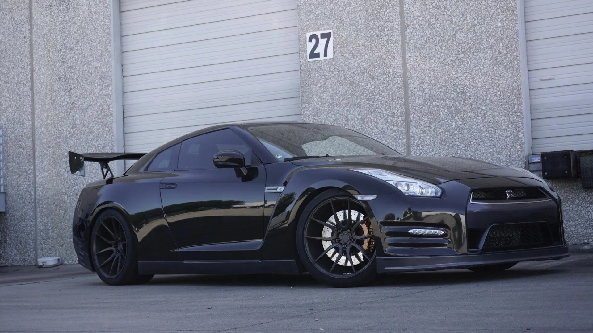 A black Nissan GT-R with aftermarket wheels and a large rear wing parked in front of a garage door.