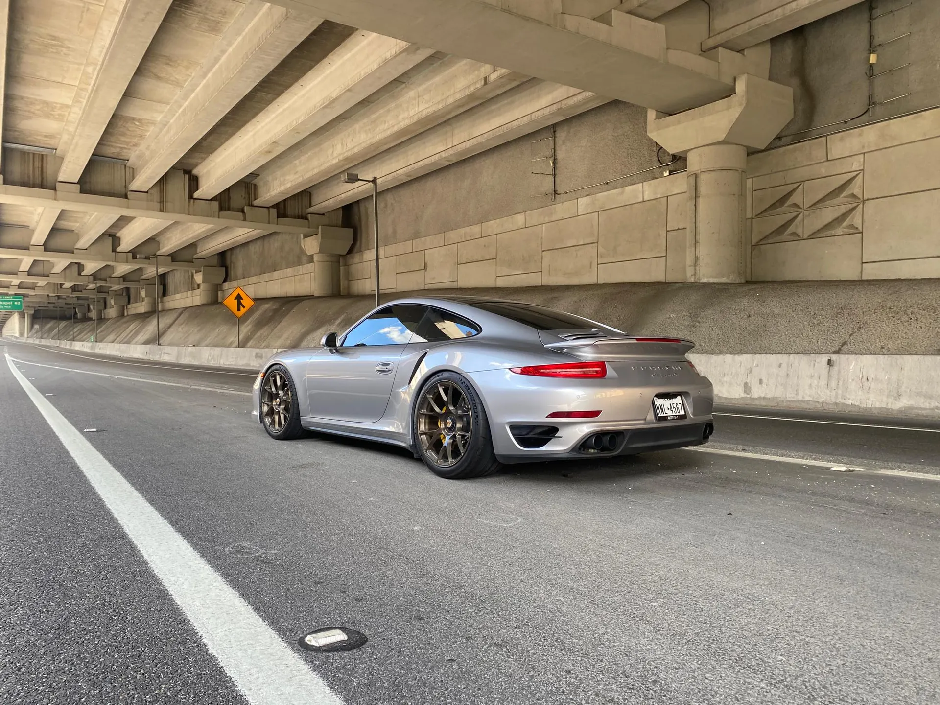 A silver Porsche 911 with custom wheels parked under a concrete overpass on a highway.