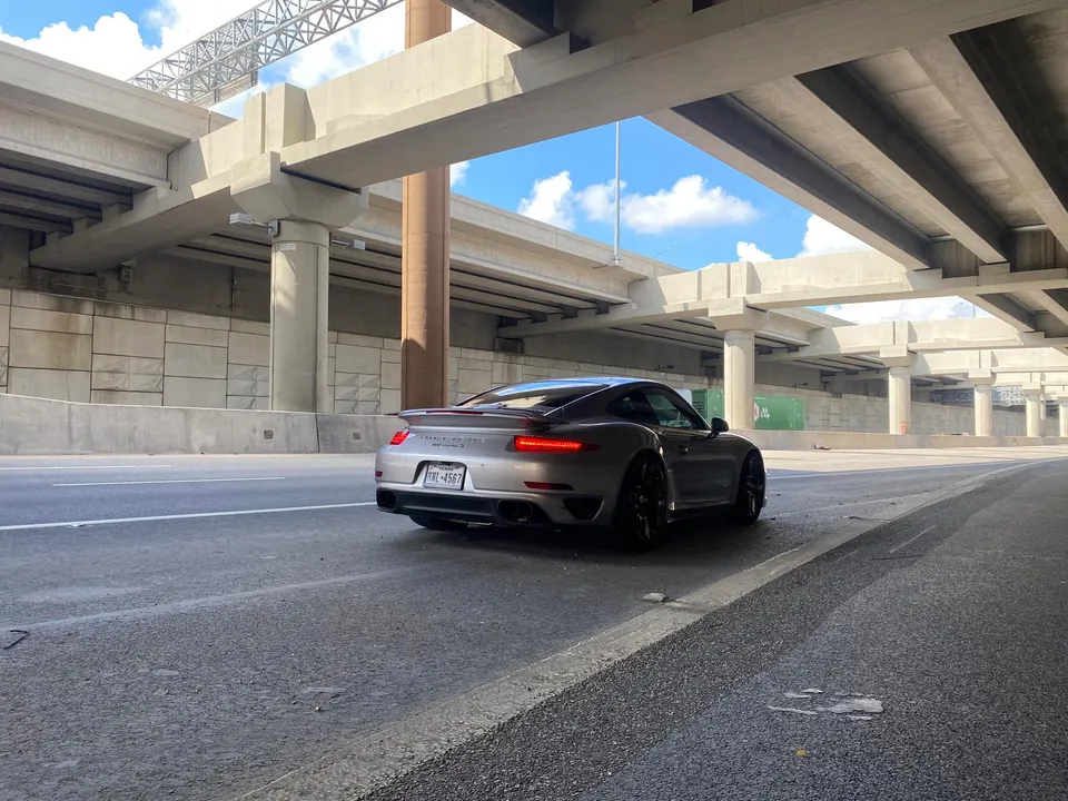 Silver Porsche 911 parked under a concrete highway overpass on a sunny day.