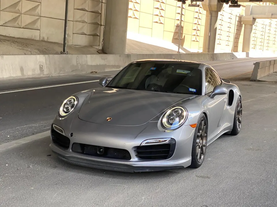 Silver Porsche 911 Turbo S parked under a concrete overpass, showcasing its sleek design and aftermarket wheels.