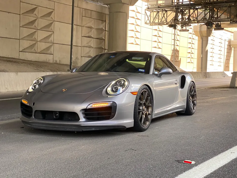 A silver Porsche 911 Turbo S with aftermarket wheels parked under a concrete overpass.