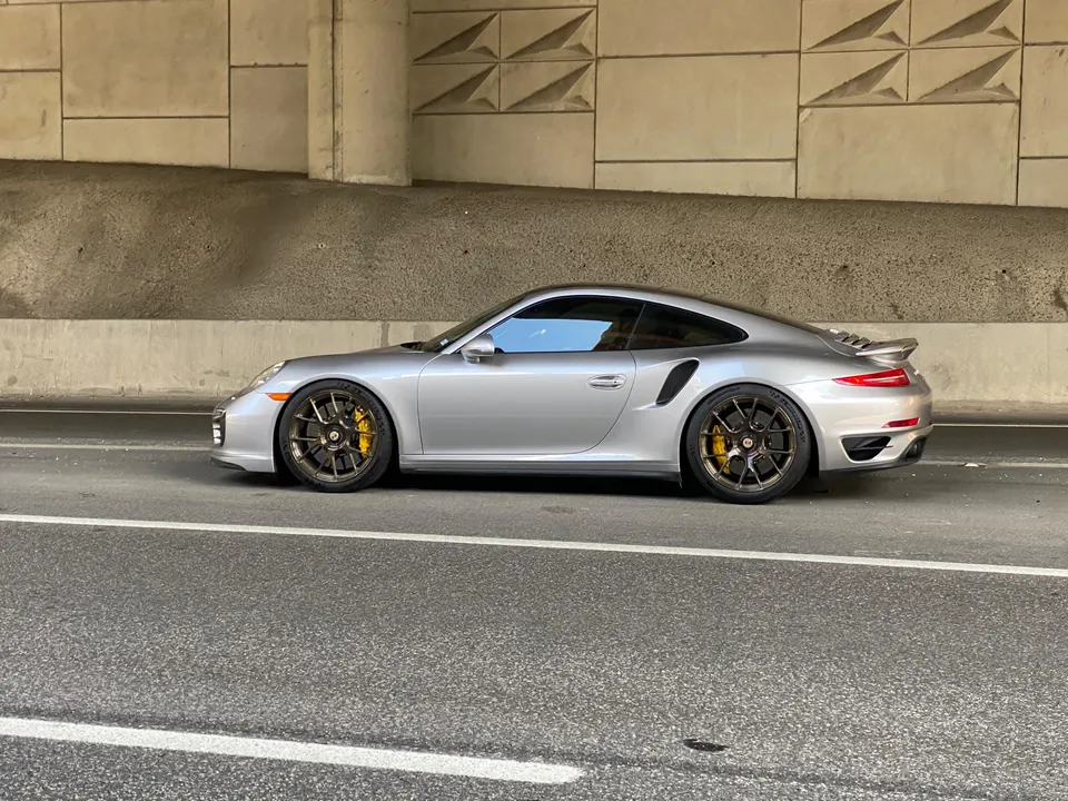 Silver Porsche 911 Turbo S with aftermarket bronze wheels and yellow brake calipers parked under an overpass.