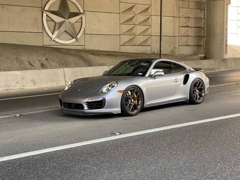 Silver Porsche 911 Turbo S parked on a road with a large Texas star emblem on the concrete wall.
