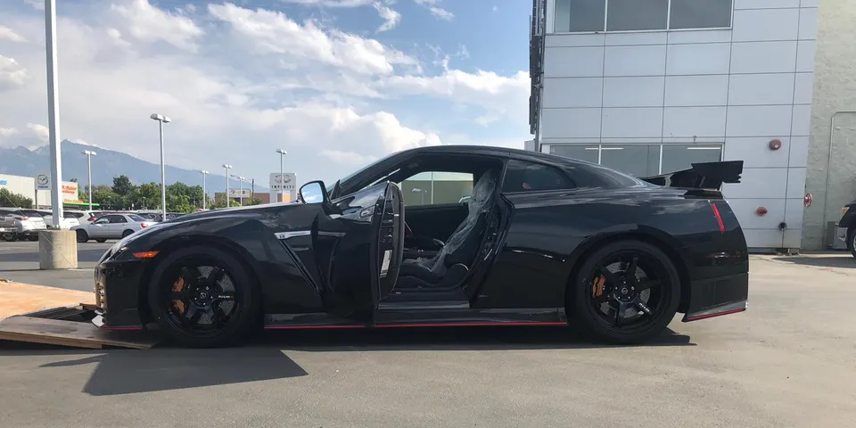 Black Nissan GT-R Nismo R35 parked in a lot with the driver's door open, showing its side profile and large rear wing.