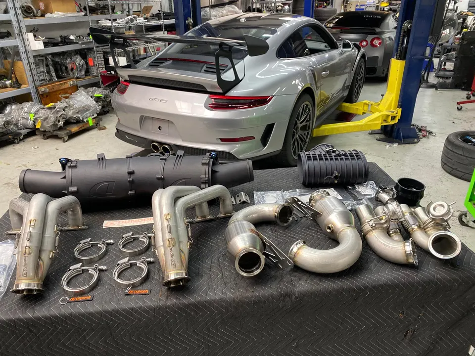 Silver Porsche GT3RS on a lift in a performance shop with Dundon Motorsports exhaust headers and an intake system on a table.