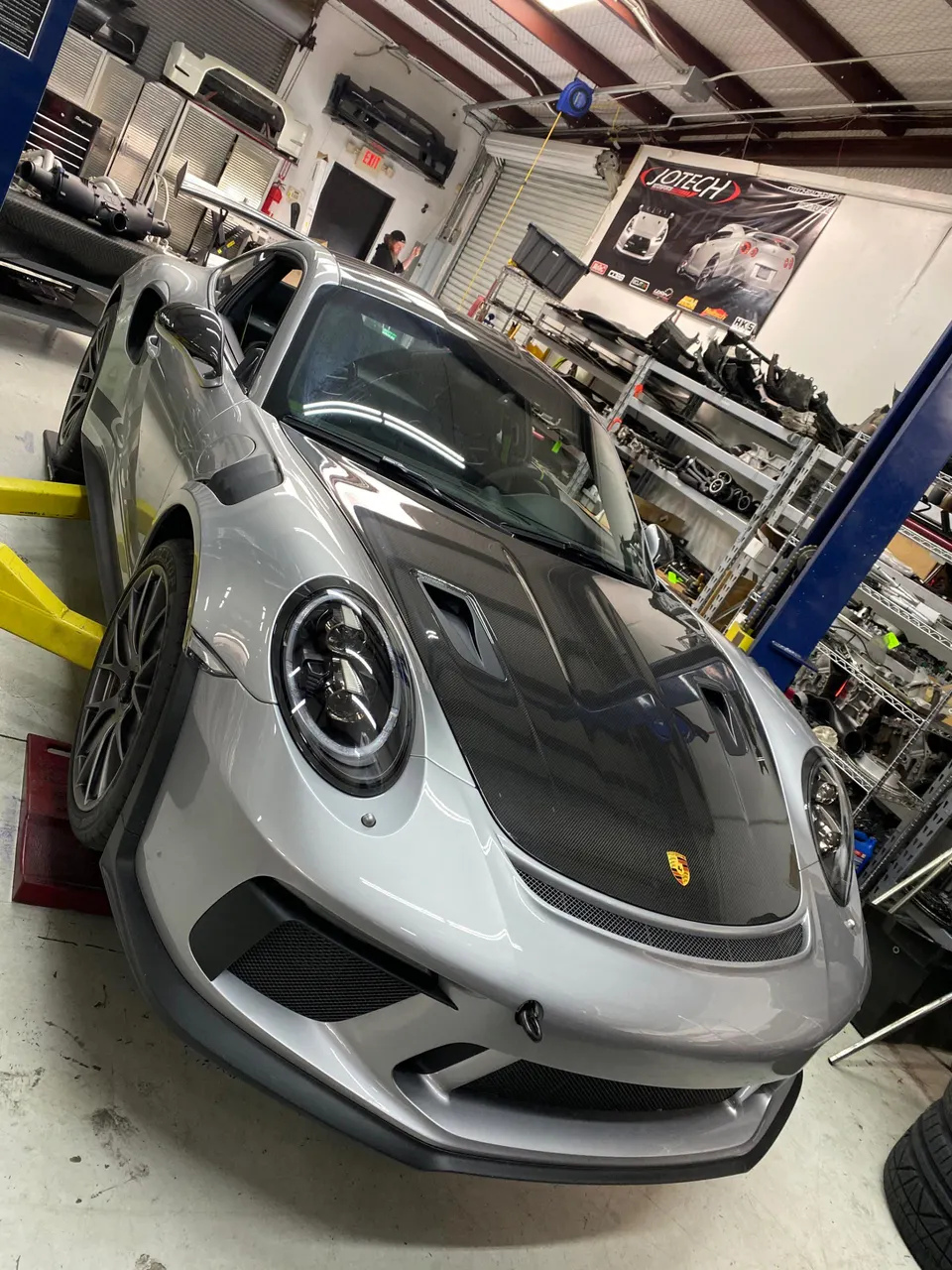 A silver Porsche GT3 RS with a carbon fiber hood on a lift in a performance tuning shop.