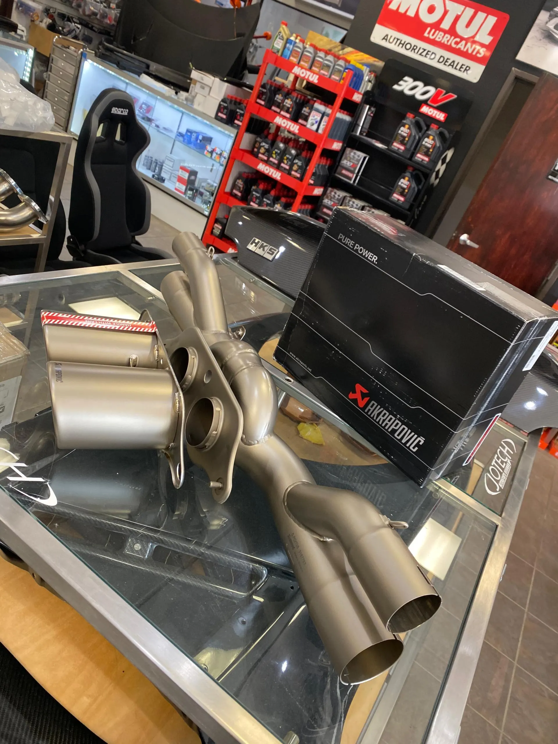 Akrapovic titanium exhaust system displayed on a glass table in a performance tuning shop with Motul lubricants and Sparco seat in background.
