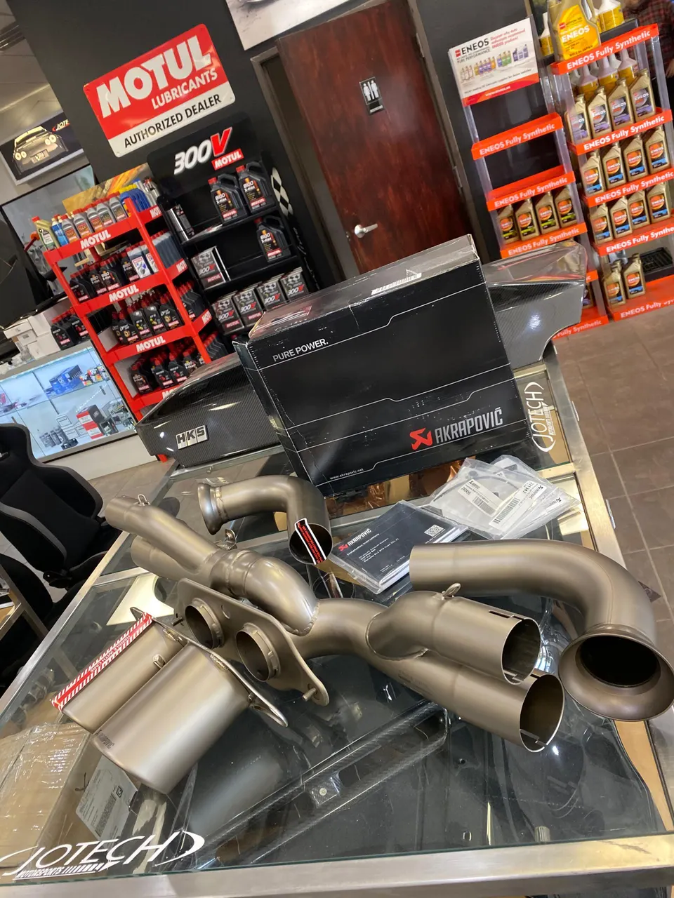 Akrapovic exhaust system and HKS carbon intake box displayed on a glass table in a performance tuning shop.
