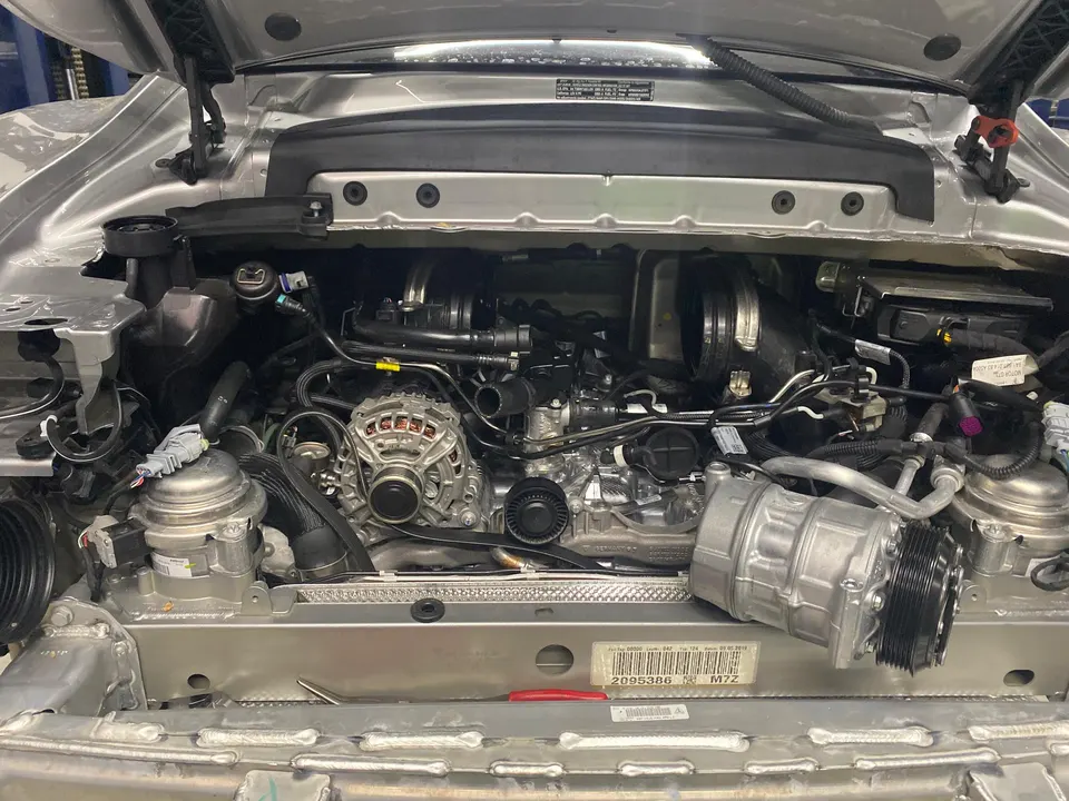 Porsche engine bay with exposed engine, alternator, and AC compressor during service at a performance shop.