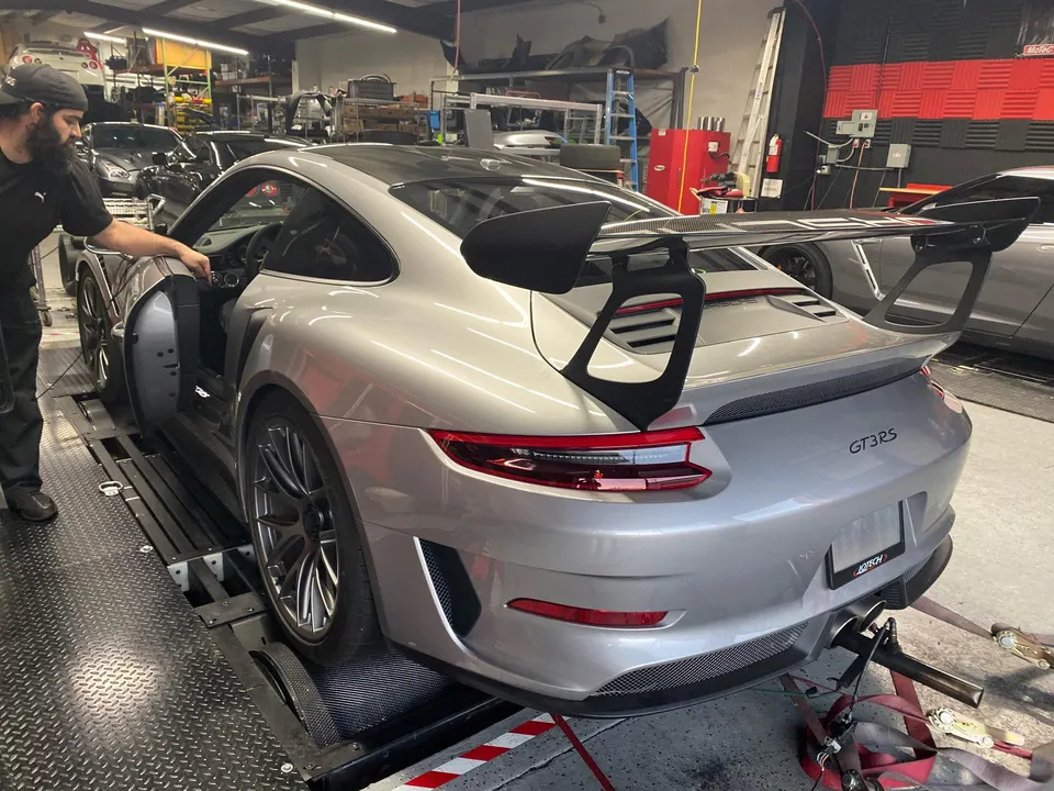 A silver Porsche GT3RS strapped to a dynamometer in a performance tuning shop, with a person standing by the driver's door.