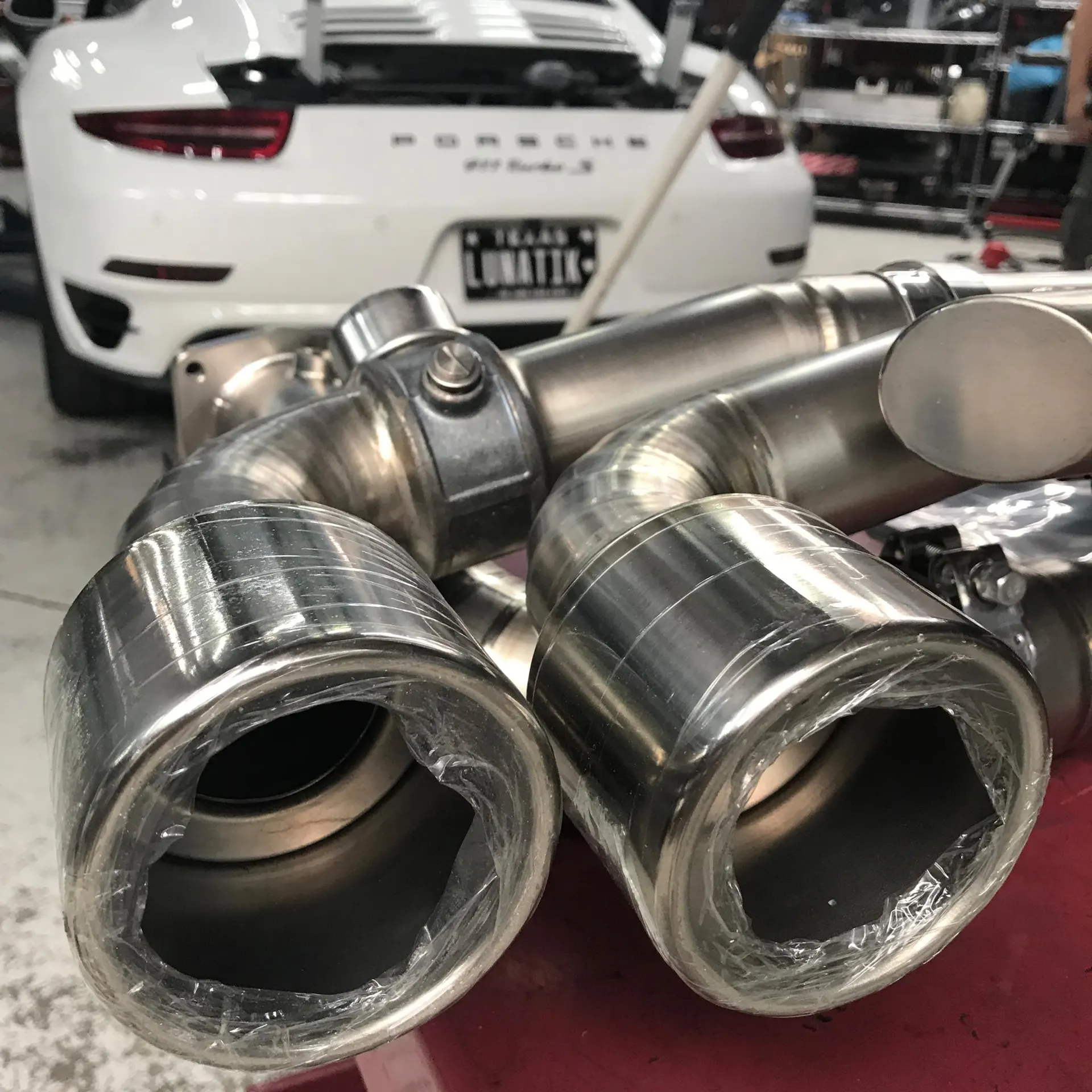 New titanium exhaust system with polished tips in the foreground, with a white Porsche 911 Turbo S in a shop background.