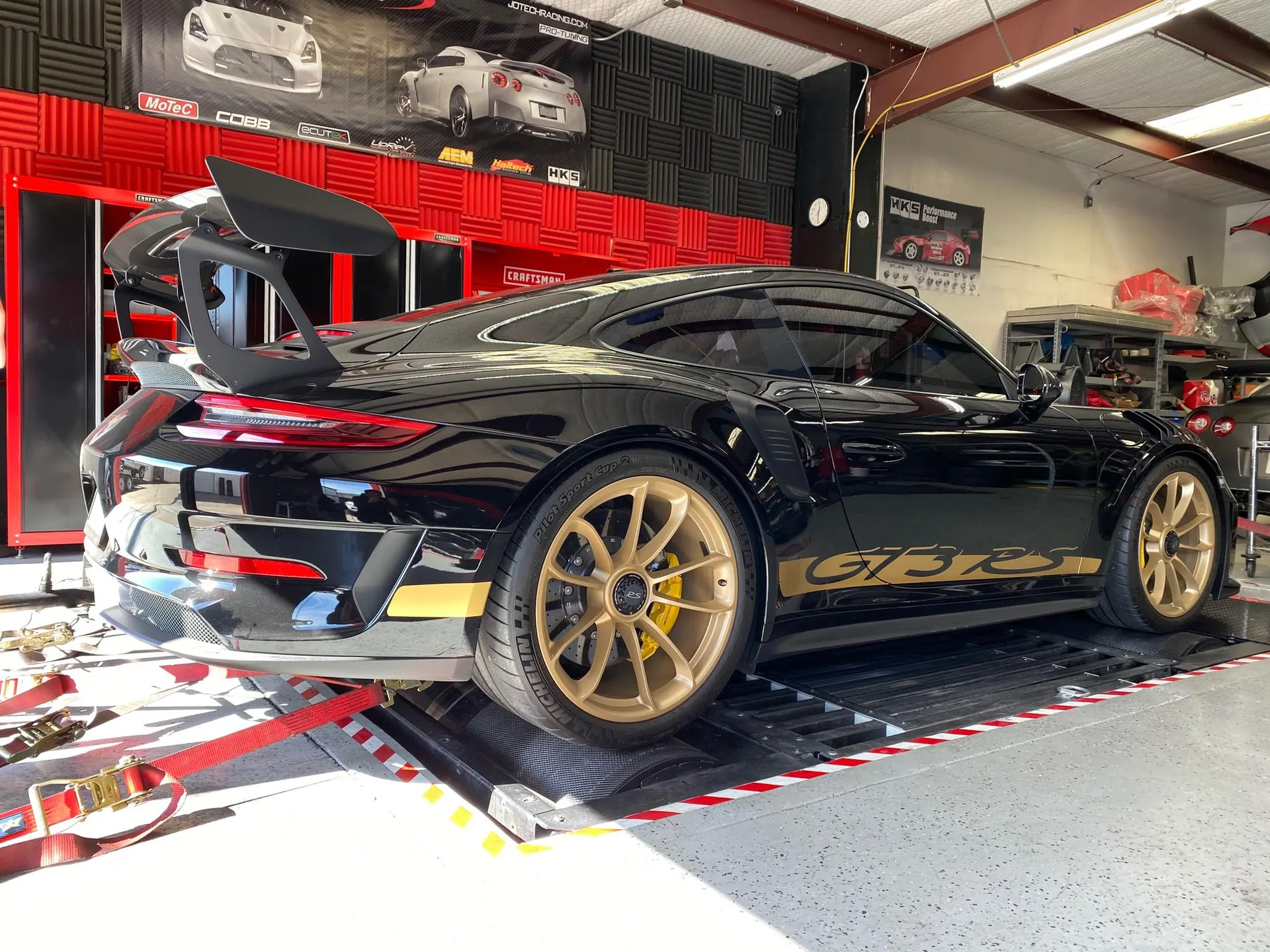 Black Porsche GT3 RS with gold wheels strapped to a dyno in a performance tuning shop.