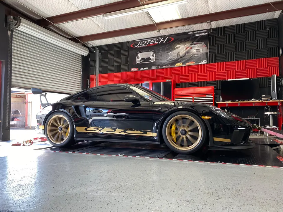 A black Porsche GT3 RS with gold wheels and accents on a dyno at Jotech Motorsports performance shop.