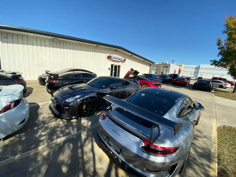 Multiple high-performance cars including Nissan GT-Rs and a Porsche GT3 RS parked outside Jotech Motorsports tuning shop.