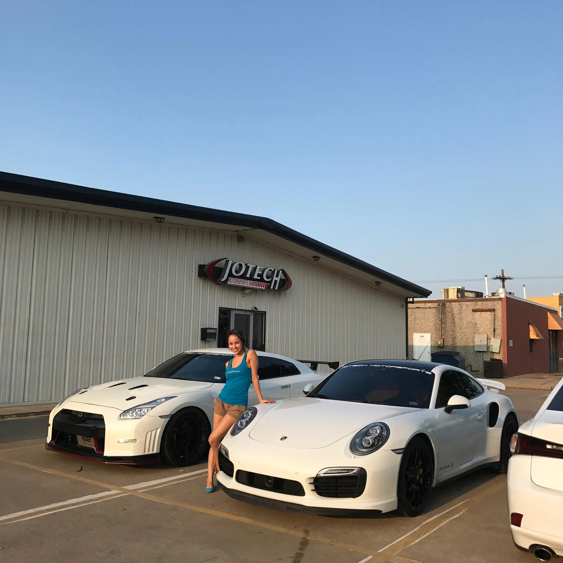 A woman poses with a white Nissan GT-R and a white Porsche 911 outside Jotech Motorsports.