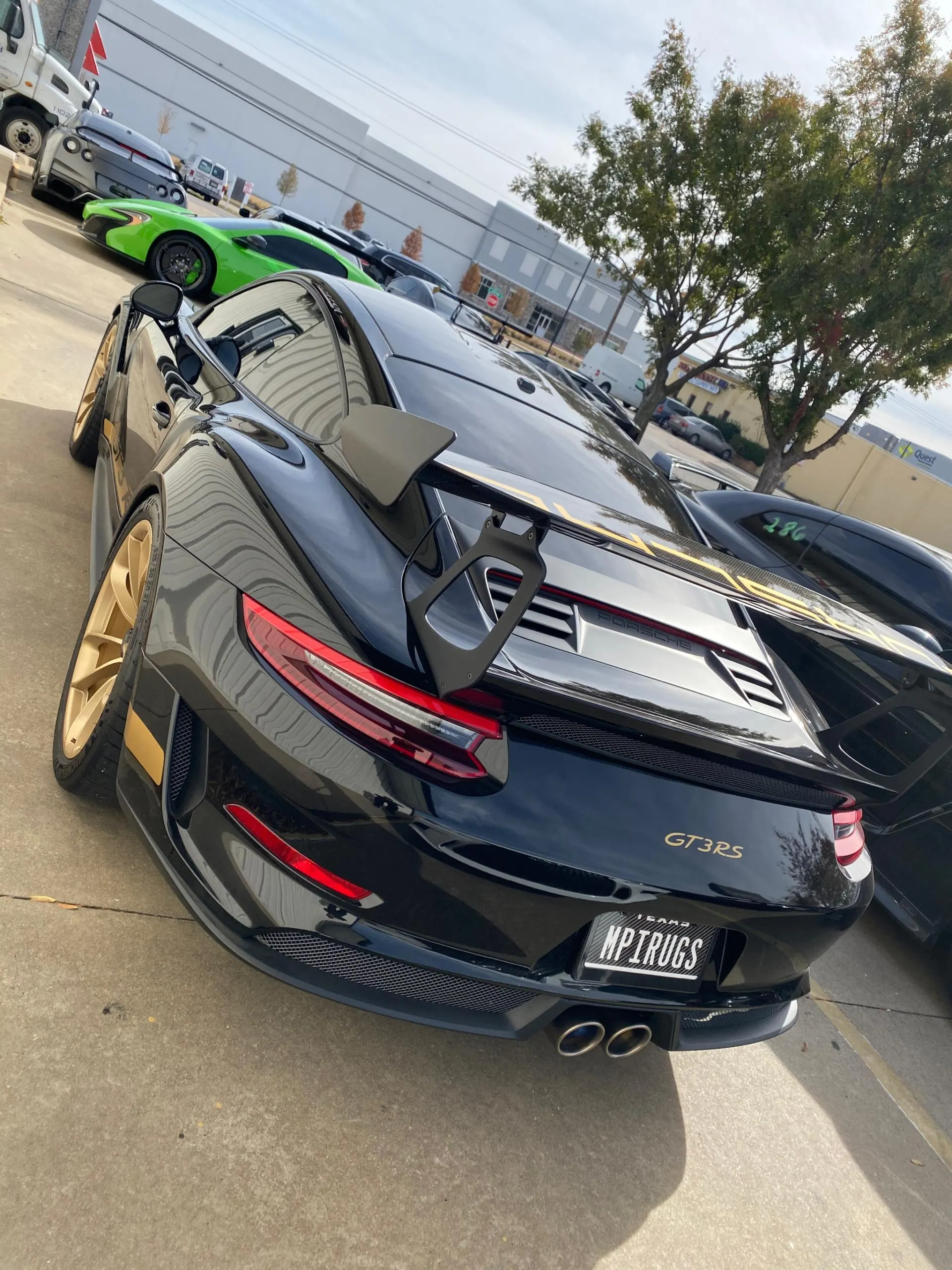 Black Porsche GT3 RS with gold wheels and a large rear wing parked outdoors, with a green McLaren in the background.