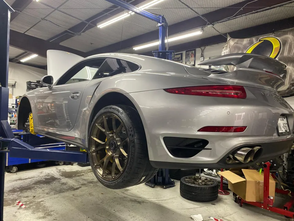 Silver Porsche 911 on a shop lift with custom bronze wheels and yellow brake calipers, undergoing service.