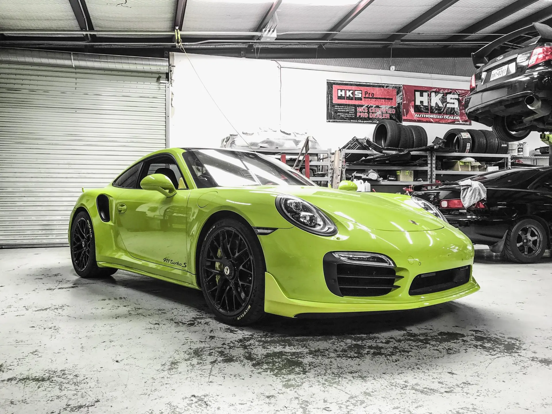 A vibrant lime green Porsche 911 Turbo S parked inside a performance tuning shop, with HKS banners in the background.