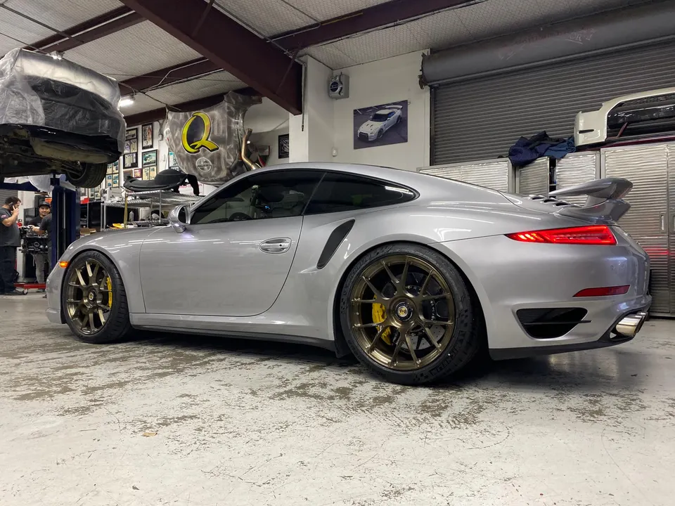 Silver Porsche 991 Turbo S with aftermarket bronze wheels and yellow brake calipers in a performance tuning shop.
