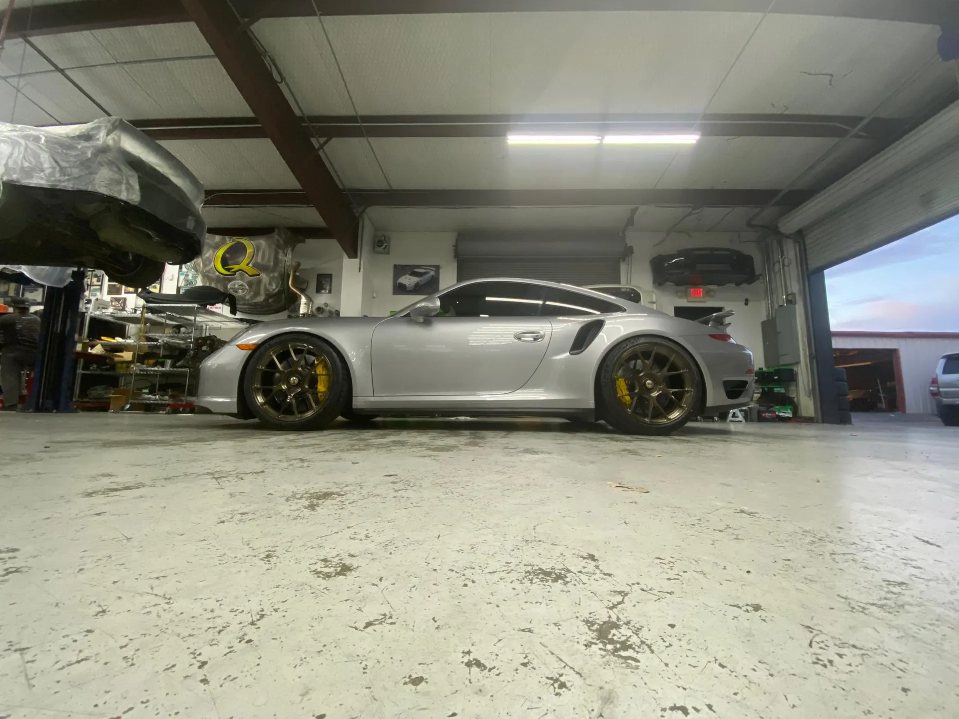 A silver Porsche 911 parked inside a performance tuning shop with another car on a lift in the background.