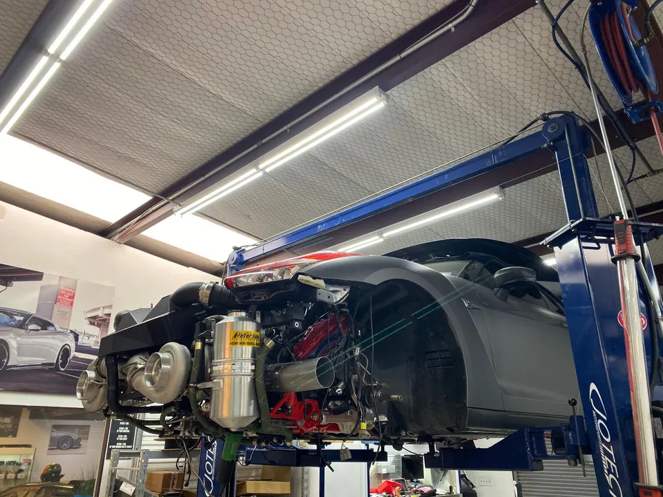 A Nissan GT-R on a lift in a performance tuning shop, with its engine bay exposed showing large twin turbochargers.
