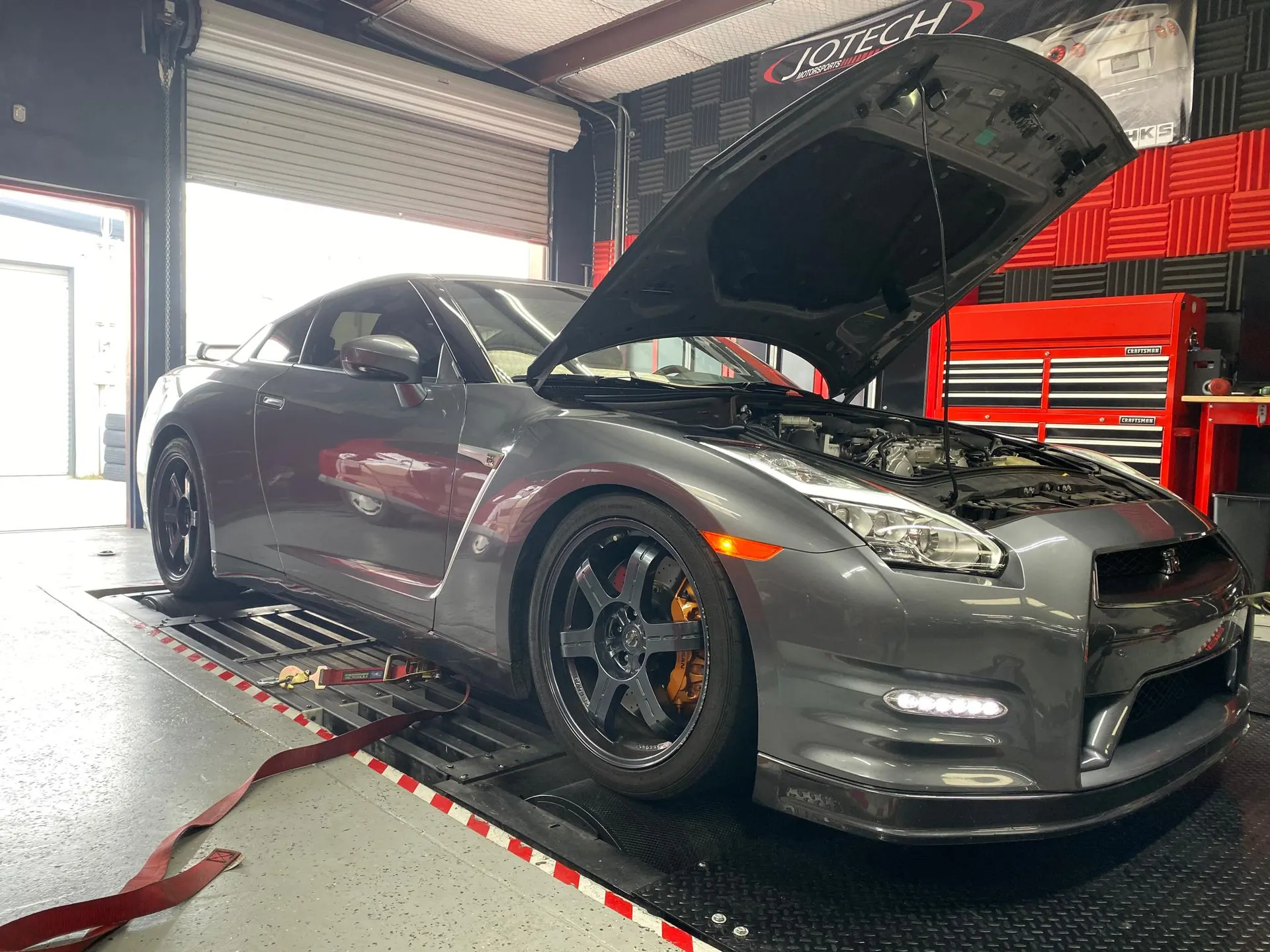 Grey Nissan GT-R with hood open strapped to a dynamometer at Jotech Motorsports for performance tuning.