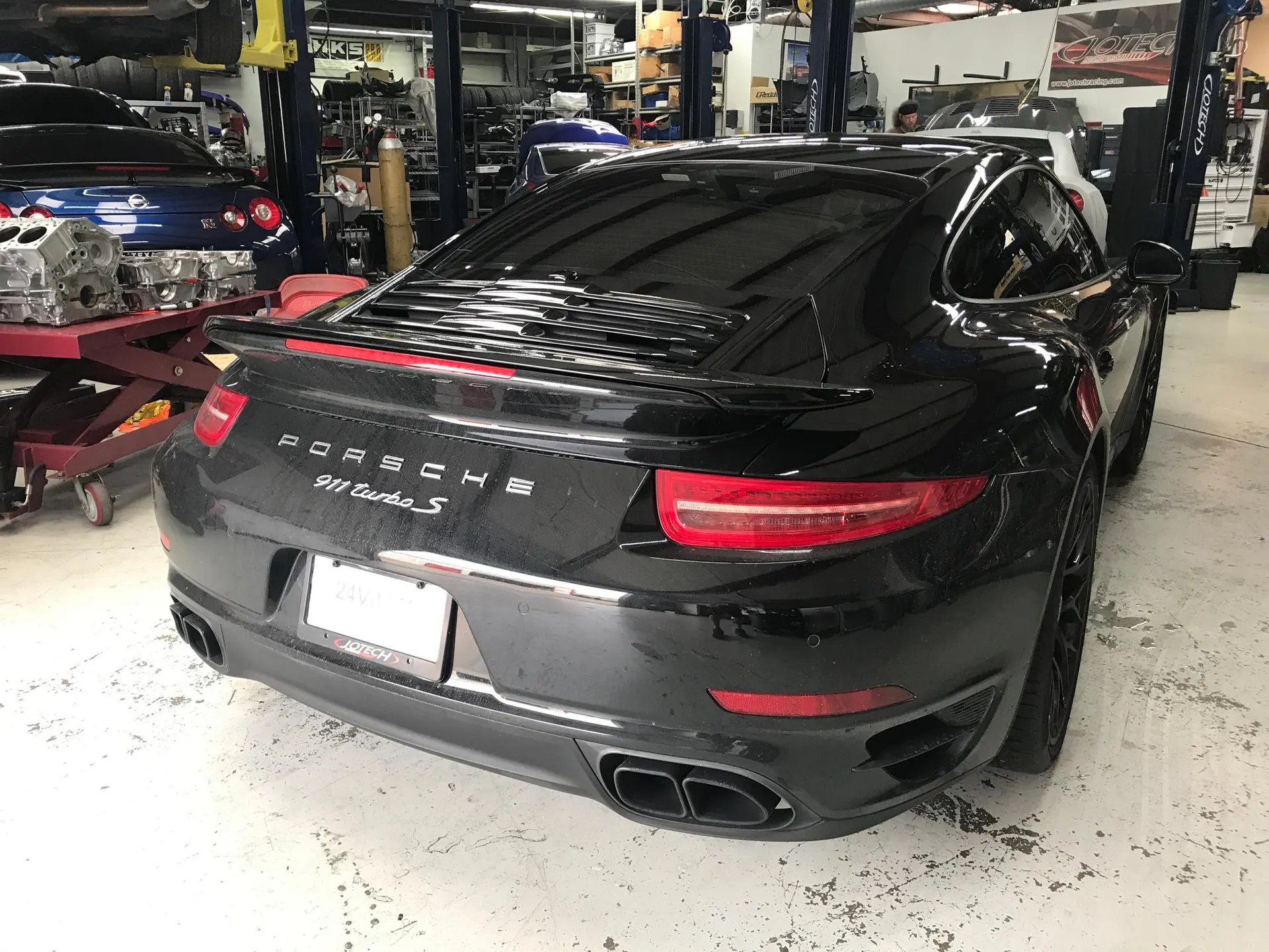 A black Porsche 911 Turbo S parked in a performance tuning shop with other cars and engine parts in the background.