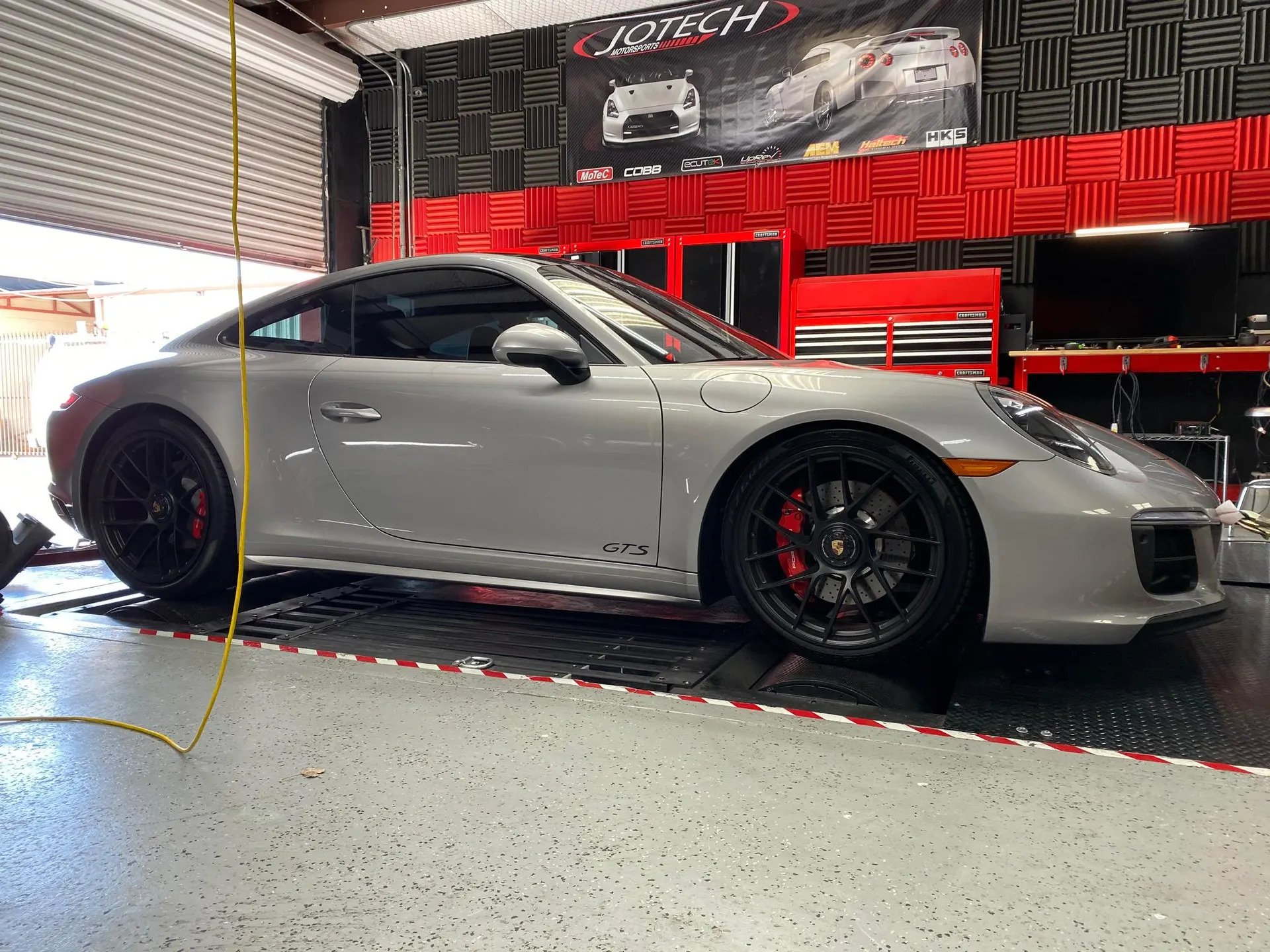 A silver Porsche 911 GTS on a dynamometer at Jotech Motorsports performance tuning shop.