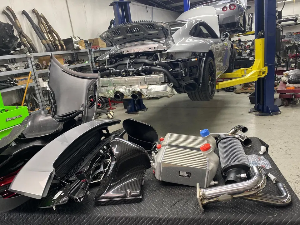 Porsche 911 on a lift in a performance shop with intercoolers, air filter, and exhaust parts laid out for installation.