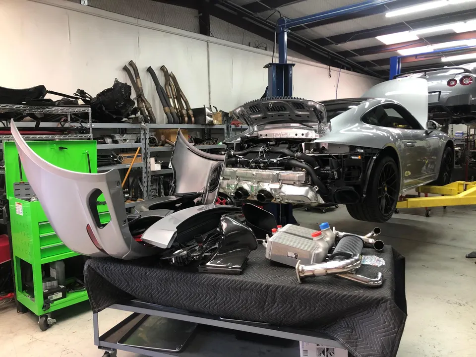 A silver Porsche 911 undergoing a performance build in a shop, with exhaust components and an intercooler on a table.