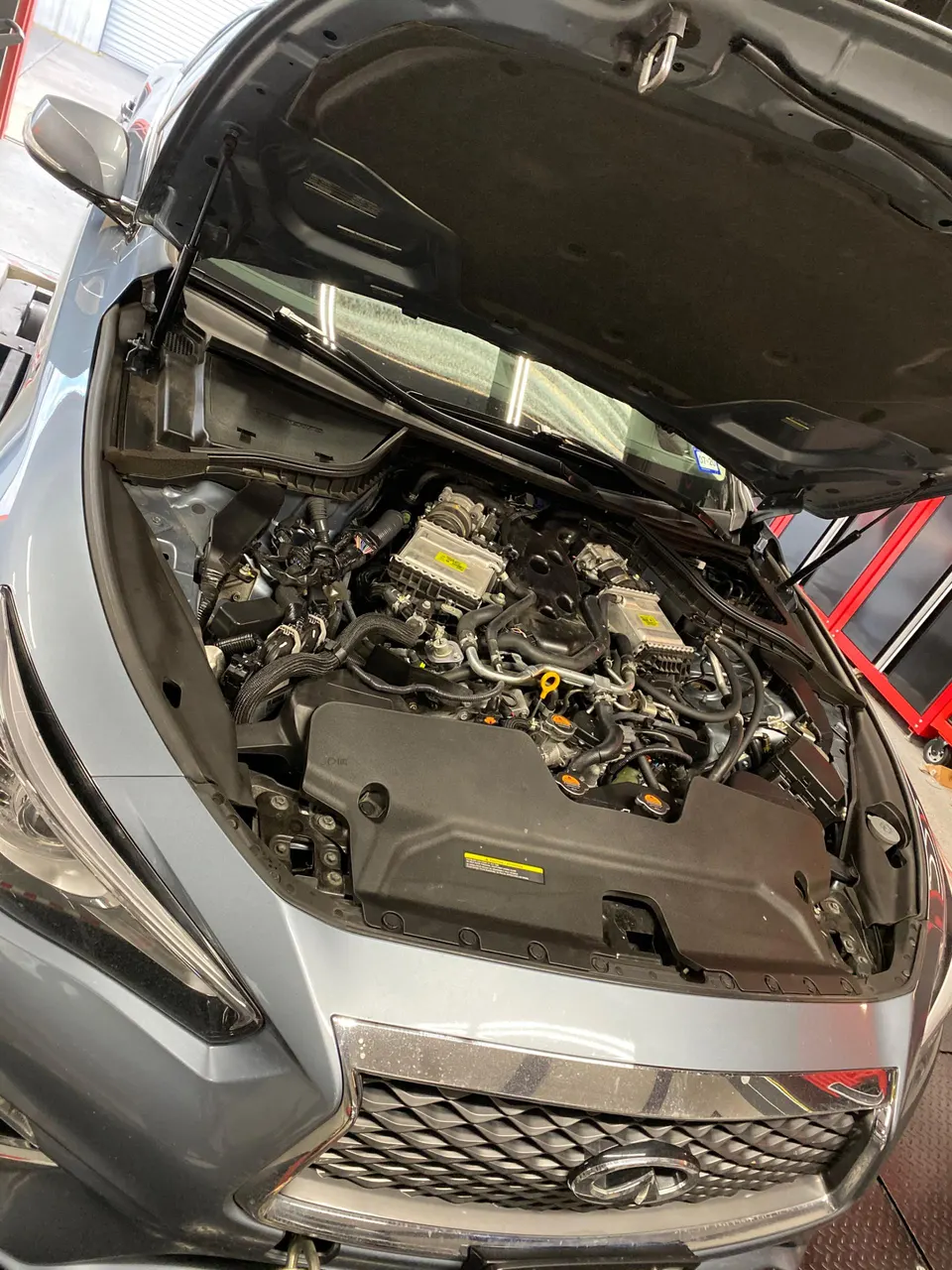 A grey Infiniti Q50 with its hood open, revealing the VR30DDTT twin-turbo engine in a shop setting.