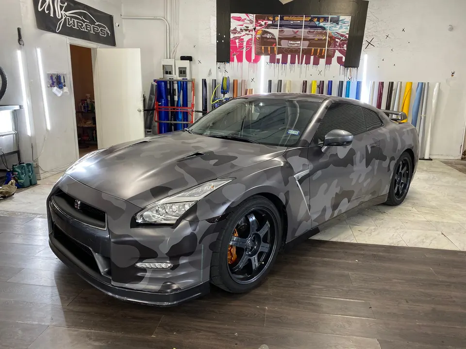 A Nissan GT-R with a grey camouflage wrap parked inside a car wrap shop with vinyl rolls in the background.