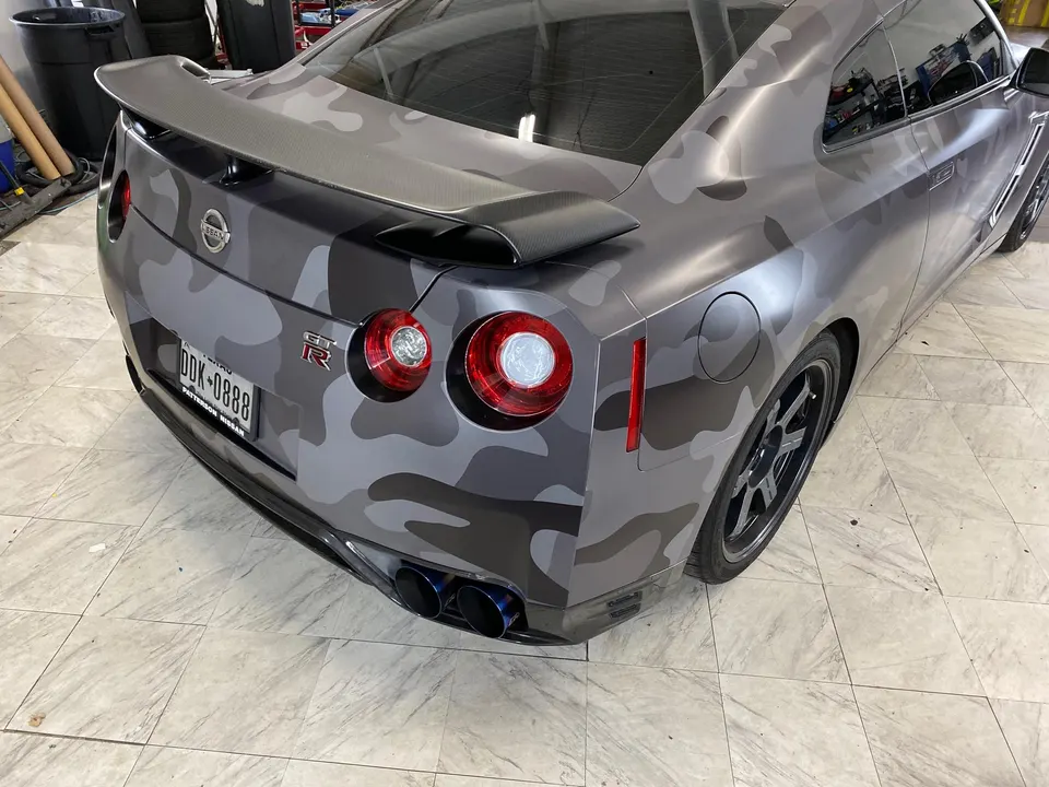 Rear view of a Nissan GT-R with a grey camouflage wrap, carbon fiber wing, and blue-tipped aftermarket exhaust.