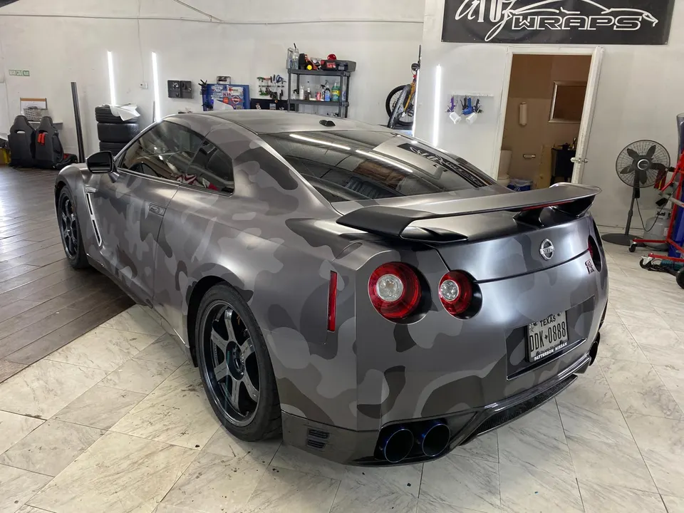 Rear three-quarter view of a Nissan GT-R with a gray camouflage wrap, aftermarket wheels, and blue-tipped exhaust in a shop.