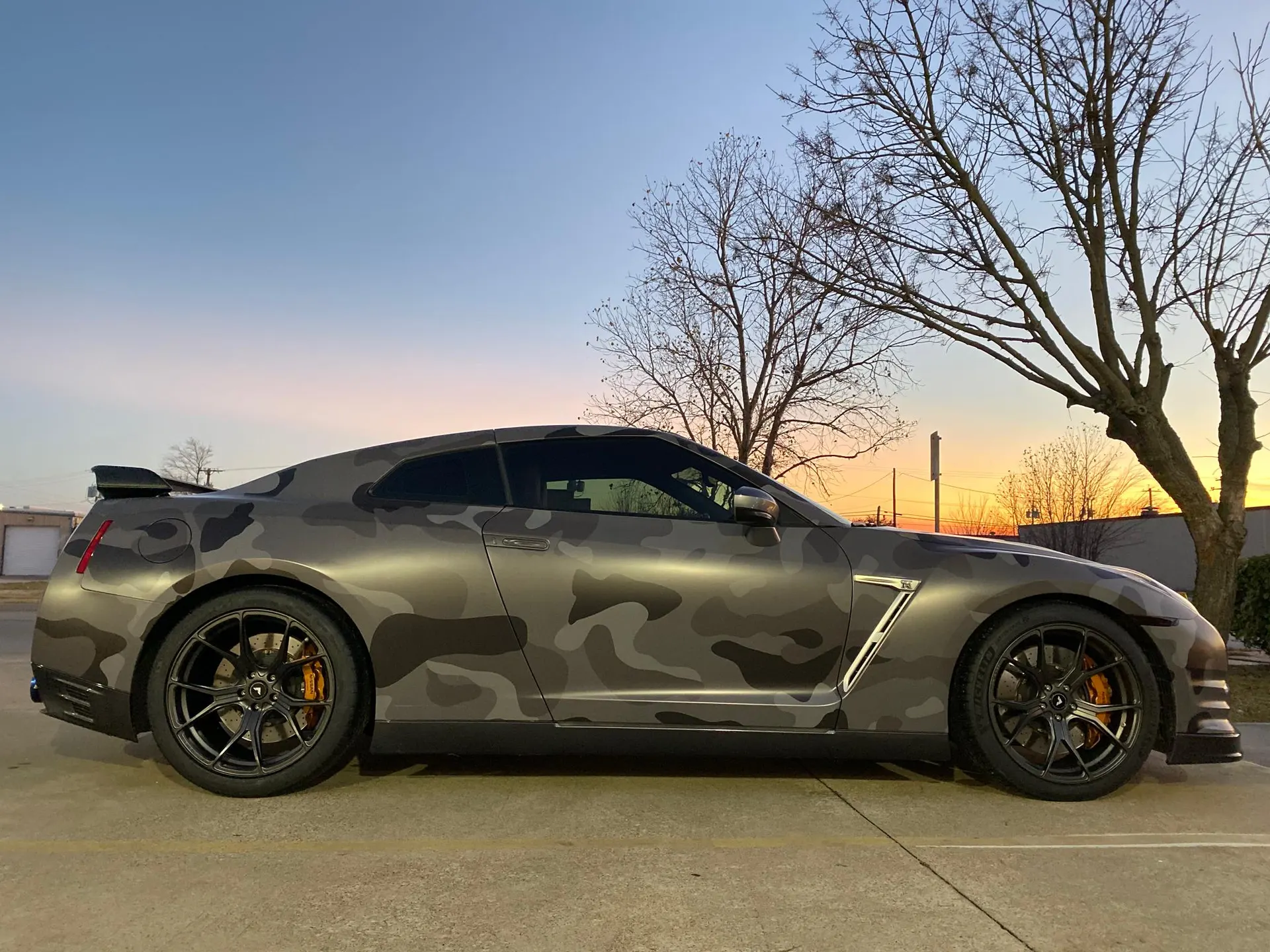 A Nissan GT-R with a camouflage wrap parked outdoors at sunset, showcasing its side profile and aftermarket wheels.