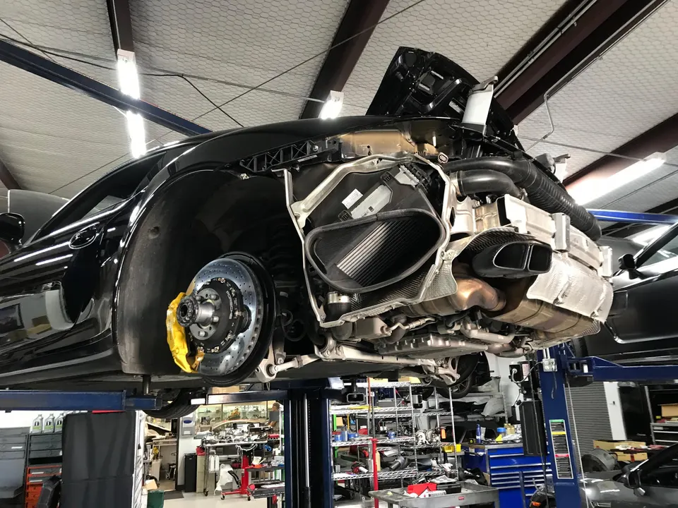 Black Porsche 911 on a lift in a shop, with the rear bumper removed exposing the exhaust system, carbon fiber air ducts, and large brake rotors.